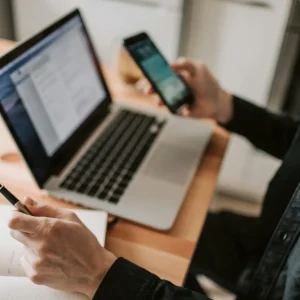 A person multitasking with a laptop and smartphone at a wooden desk.